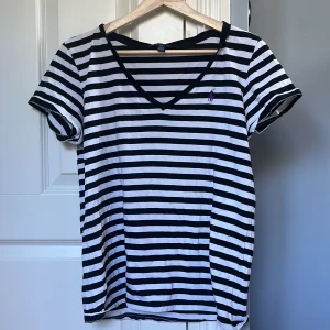 Randig t-shirt från Ralph Lauren - Snygg t-shirt från Ralph Lauren med svartvita ränder och lila broderad logga på bröstet. T-shirten har v-ringning, korta ärmar och är gjord i mjuk bomull. Perfekt för en chill och stilren look.