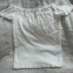 Vit ribbad offshoulder topp - Supersöt vit ribbad topp med offshoulder-modell och korta ärmar. Den har en lätt rynkad kant upptill och nertill för en extra gullig touch. Perfekt för varma dagar och enkel att styla med jeans eller kjol.