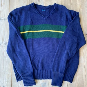 Blå v-ringad tröja från Polo Ralph Lauren - Säljer en blå v-ringad tröja från Polo Ralph Lauren i 100% bomull. Tröjan har grönt och gult randigt band över bröstet och den klassiska broderade loggan på vänster sida. Ribbstickade muddar och avslappnad passform. Perfekt för dig som gillar preppy stil.