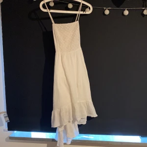 Vit sommarklänning med volang - Superfin vit klänning från BINDEX med smala axelband, broderad överdel och volangig nederkant. Lätt och luftig modell, perfekt för varma dagar. Sitter snyggt med sin asymmetriska fåll och romantiska känsla.