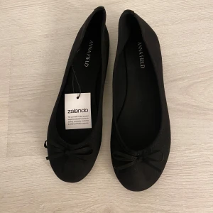 Svarta ballerinaskor från Anna Field - Klassiska svarta ballerinaskor från Anna Field med rund tå och söt rosettdetalj framtill. Skorna har en platt sula och är tillverkade i ett mjukt tygmaterial som känns bekvämt på foten. Perfekta för en stilren och enkel look.