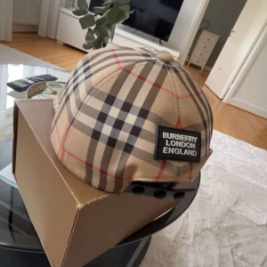 Burberry rutig keps beige M - Snygg keps från Burberry i klassiskt beige rutigt mönster med svarta, vita och röda linjer. Justerbar baktill med svart spänne och tydlig Burberry London England-patch. Tillverkad i bomull och polyester, perfekt för dig som vill sticka ut med stil.