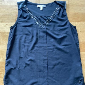 Svart linne med pärldetaljer Esprit - Svart linne Esprit med snygga pärldetaljer längs v-ringningen och axelparti i skinnimitation. Linnet har en rak passform och är perfekt för dig som gillar stilrena plagg med en twist.