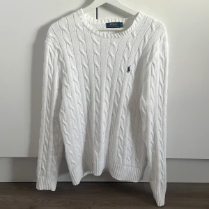 Vit kabelstickad tröja Polo Ralph Lauren - Vit kabelstickad tröja från Polo Ralph Lauren med klassisk rund halsringning och broderad marinblå logga på bröstet. Tröjan har ribbade muddar vid ärmslut och nederkant, perfekt för en clean och stilren look.