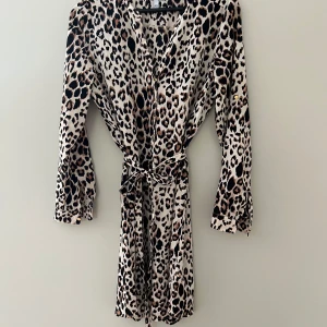 Leopardmönstrad kortklänning KappAhl - Snygg kortklänning från KappAhl med leopardmönster i beige, svart och brunt. Klänningen har långa ärmar, knappar framtill, v-ringning och ett matchande knytbälte i midjan. Perfekt för dig som vill sticka ut med ett djurmönstrat plagg.