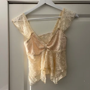 Beige spetsig blus med volang - Romantisk blus i ljusbeige spets med breda axelband och v-ringning. Toppen har volangdetaljer framtill och är helt genombruten med blommigt spetsmönster. Perfekt för dig som gillar en feminin och drömmig stil.