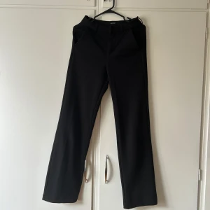 Svarta kostymbyxor från Vero Moda - Stilrena svarta kostymbyxor från Vero Moda med raka ben och klassisk passform. Byxorna har hällor för bälte och sidfickor, perfekta för en clean och snygg look. Materialet är mjukt och följsamt, vilket gör dem bekväma hela dagen.