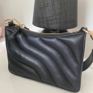 Svart axelväska med gulddetaljer och i bra skick - Snygg svart axelväska i quiltad design med vågiga sömmar. Väskan har bred axelrem och dragkedja i guld. Perfekt storlek för det viktigaste och passar till många outfits. Väldigt bra skick