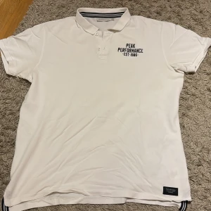 Vit Peak Performance pikétröja XXL - Snygg vit pikétröja från Peak Performance i storlek XXL. Klassisk krage med knappar och svart logga på bröstet. Tillverkad i bomull, perfekt för en clean och sportig look. Passar dig som gillar stilrena plagg med en sportig touch.