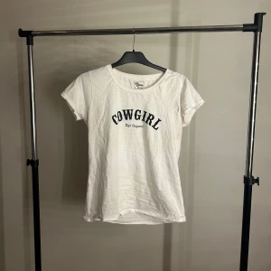 Vit COWGIRL t-shirt High Chaparral - Vit t-shirt från High Chaparral med svart COWGIRL-tryck på bröstet. Klassisk rund halsringning och korta ärmar. Tillverkad i 100% bomull för en skön och avslappnad känsla. Perfekt för dig som gillar western vibes och enkel stil.