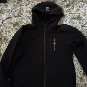 Svart zip hoodie från Sail Racing - Svart zip hoodie från Sail Racing med huva och hel dragkedja framtill. En bröstficka med dragkedja och broderad logga på bröstet samt på huvan.