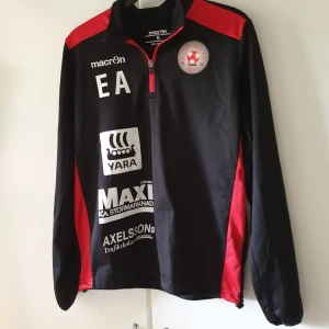 Svart och röd träningströja Macron S - Sportig svart och röd långärmad träningströja från Macron med halv dragkedja och tryckta loggor från Köping FF och sponsorer. Perfekt för fotbollsträning, med bekväm passform och snygga röda detaljer på axlar och sidor. Står initialer EA på den. Tröjan har tidigare befunnit sig I samma hem som en katt. Kan eventuellt råka följa med något enstaka katthår.