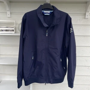 Marinblå vindjacka från Sebago - Snygg marinblå vindjacka från Sebago med dragkedja framtill och två fickor med zip. Jackan har ståkrage och tryck på ärmen med texten 'Sebago Marine Sailing Crew'. Perfekt för dig som gillar stilren och sportig look. Använd endast en gång. Nypris 2100:- 
