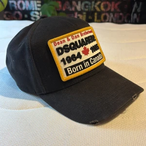 Svart keps från Dsquared2 med patch - Svart keps från Dsquared2 med stor broderad patch framtill i gult, vitt, rött och svart med texten 'Born in Canada' och lönnlöv. Justerbar rem bak med metallspänne och broderad text 'Family Business'. Klassisk böjd skärm och cool streetkänsla.