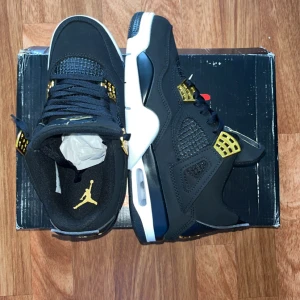 Nike Air Jordan 4 Retro Black/Gold - Säljer ett par Nike Air Jordan 4 Retro i svart med guldiga detaljer och vit sula. Skorna har klassisk meshpanel, snörning och Jumpman-logga i guld på innersulan. Stilren och ikonisk siluett som sticker ut med sina kontrasterande färger.