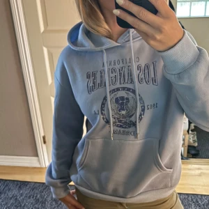 Ljusblå hoodie med Los Angeles-tryck - Ljusblå hoodie med tryck framtill där det står 'California Los Angeles 1982 Dream'. Tröjan har huva med snörning, magficka och ribbade muddar. Mjuk insida och normal passform.