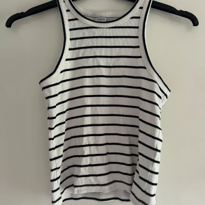 Randig ribbad linnetopp från Lindex - Snygg vit ribbad topp från Lindex med svarta horisontella ränder. Toppen har ärmlös design och rund halsringning, perfekt för en avslappnad och trendig look. Materialet är mjukt och stretchigt för extra komfort.