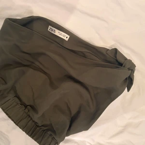 Olivgrön bandeau tubtopp från Zara - Säljer en olivgrön bandeau tubtopp från Zara med snygg knytdetalj på ena sidan. Toppen är ärmlös och har resår nedtill för en bekväm passform. Perfekt till varma dagar och enkel att styla med jeans eller kjol.