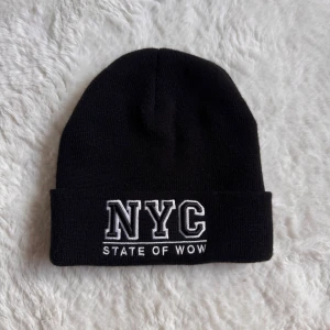 Svart NYC mössa - Svart ribbstickad mössa med uppvikt kant och vit broderad text 'NYC STATE OF WOW' framtill. Enkel och stilren design som passar till det mesta. Perfekt accessoar för kalla dagar.