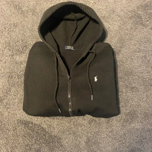 Svart hoodie från Polo Ralph Lauren - Svart hoodie från Polo Ralph Lauren med dragkedja och huva. Klassisk broderad logga på bröstet och justerbara snören vid huvan. Inte äkta .Mjuk och skön, perfekt för chill dagar. Snygg och enkel att matcha med allt. Storlek xs 