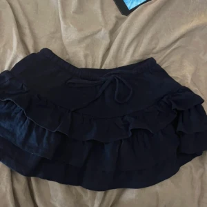 Svart volangkjol med knytband - Supersöt svart kortkjol med dubbla volanger och knytband i midjan. Perfekt för dig som gillar en lekfull och trendig stil. Kjolen har resår i midjan för extra komfort och är lätt att matcha med både sneakers och boots.