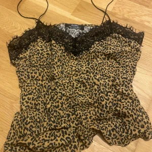 Leopardmönstrad linnetopp från Bershka - Snygg leopardmönstrad linnetopp från Bershka med tunna axelband och svart spetsdetalj vid urringningen. Toppen har en beige och svart färgkombination och är perfekt för dig som vill sticka ut med ett djurmönster. Lätt och luftig känsla.