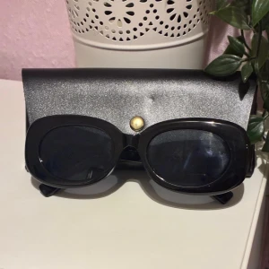 Svarta solglasögon med fodral - Snygga svarta solglasögon med breda, ovala bågar och mörka glas. Kommer med ett stilrent svart fodral med guldfärgad knapp. Perfekta för att ge en cool vibe till din outfit.
