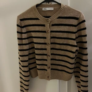 Beige och svart randig kofta från Zara - Säljer en snygg beige kofta från Zara med svarta ränder. Koftan har långa ärmar och knappar framtill. 