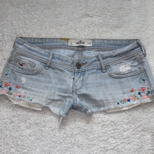 Lågmidjade Hollister shorts - Superfina ljusblå jeansshorts från Hollister med broderade blommor i flera färger längs sidorna. Shorts med fransig kant, låg midja och slitna detaljer både fram och bak. Obs, en liten orange fläck vid midjan som jag ej lyckas få bort, men syns inte så mycket när man har på sig dom. 
