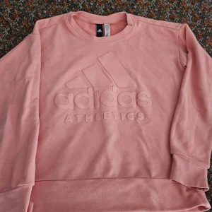 Rosa sweatshirt från Adidas - Säljer en snygg rosa sweatshirt från Adidas med deras logga och texten 'ATHLETICS' i relief på framsidan. Tröjan har långa ärmar och en rund halsringning. Perfekt för en sportig look eller en avslappnad dag.