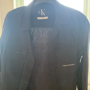 Svart skjorta från Calvin Klein Jeans - Säljer en svart skjorta från Calvin Klein Jeans i regular fit. Skjortan har klassisk krage, två bröstfickor med knappar och diskret logga broderad på ena fickan. Perfekt för dig som gillar stilrena plagg med snygga detaljer.