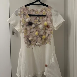 Vit blommig t-shirt med 3D-blommor - Superfin vit t-shirt med blommigt mönster och 3D-blommor i olika färger på bröstet. T-shirten har rund halsringning och korta ärmar. Perfekt för dig som vill sticka ut med en unik och söt topp!