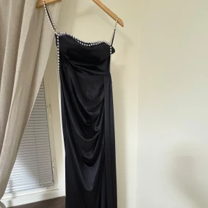 Svart långklänning med pärlband från SHEIN - Elegant svart långklänning med smala axelband dekorerade med pärlor. Klänningen har figurnära passform och draperad effekt framtill. Perfekt för festliga tillfällen när du vill sticka ut. Aldrig använd helt ny.