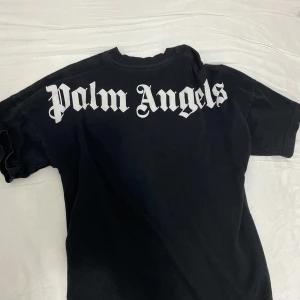 Svart t-shirt från Palm Angels - Säljer en svart t-shirt från Palm Angels med stor vit logga på ryggen och mindre logga i nacken. Klassisk passform och tillverkad i 100% bomull. Perfekt för dig som gillar streetwear och vill sticka ut med en ikonisk look. T-shirten är ganska tjock så kan även användas på sämre väder 