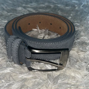 Blå mockabälte med silverfärgat spänne - Snyggt blå bälte i mocka med klassiskt silverfärgat metallspänne. Bältet har stilrena sömmar längs kanterna och insidan är brun. Perfekt accessoar för att lyfta din outfit.