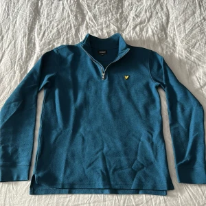 Blå långärmad tröja med dragkedja från Lyle & Scott - Säljer en blå långärmad tröja från Lyle & Scott Junior med halv dragkedja och klassisk krage. Den passar bra som S. Tröjan har en gul broderad logga på bröstet och är perfekt för en stilren vardagslook.