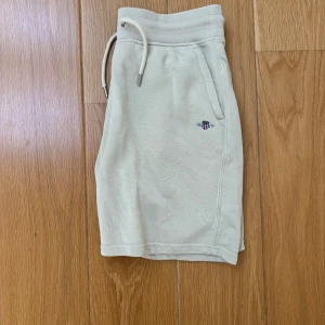 Beiga shorts från GANT med snörning - Snygga beiga shorts från GANT med ribbad midja och snörning. Shortsen har fickor på sidorna och en liten broderad GANT-logga framtill. Perfekta för varma dagar och enkel att matcha med olika toppar.