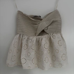 Beige croppad linnetopp med spetsdetaljer. liten i storleken, passar S och XS - Supersöt croppad topp med virkad beige överdel och broderad, vit spets nertill. Smala axelband och omlott-detalj framtill. Perfekt för varma dagar och ger en romantisk vibe.