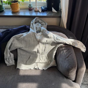 Grå och blå hoddie från Polo Ralph Lauren - Grå hoodie från Polo Ralph Lauren med dragkedja och huva. Klassisk design med två fickor framtill och liten broderad logga på bröstet. Perfekt för en avslappnad stil.