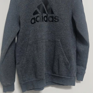 Grå hoodie från Adidas - Säljer en stilren grå hoodie från Adidas med deras ikoniska logga i svart på bröstet. Tröjan har en stor ficka framtill och en bekväm huva. Perfekt för en avslappnad stil.