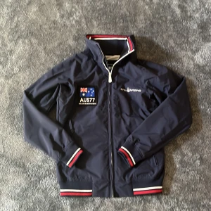 Marinblå Sail racing jacka  - Sällsynt och snygg marinblå jacka från Sail Racing med broderad australiensisk flagga och texten "AUS77". Jackan har dragkedja och ribbade muddar med röd, vit och blå rand. Perfekt för en sportig look! Priset är självklart förhandlingsbart. 😊 Tveka inte att höra av dig! 🫡
