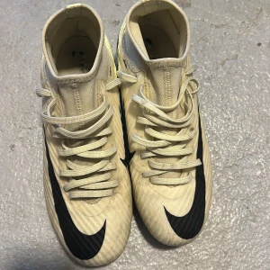 Beige fotbollsskor från Nike - Säljer ett par beige fotbollsskor från Nike med svarta detaljer och snörning. Skorna har en rund tå och är designade för optimal prestanda på planen. Perfekta för fotbollsspelare som vill ha stil och funktionalitet.