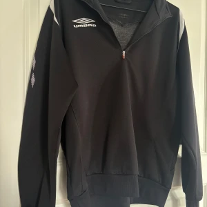 Svart träningströja med dragkedja från Umbro - Svart långärmad träningströja från Umbro med vit logga på bröstet och axlarna. Tröjan har halv dragkedja framtill och ribbade muddar. Perfekt för sport eller träning.