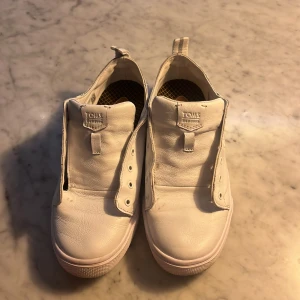 Vita sneakers från Toms - Säljer ett par vita sneakers från Toms i skinn. Skorna har låg modell men saknar skosnören. Perfekta för en clean och stilren look. Passar till det mesta och är enkla att ta på tack vare dragflik bak.
