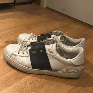 Valentino sneakers med mörkblå detalj - Säljer ett par vita Valentino sneakers med bred mörkblå rem över vristen och klassisk logga på plösen. Skorna har rund tå, snörning och en chunky sula med nitar baktill. Perfekta för dig som gillar stilrena men ändå unika sneakers. I bra skick.