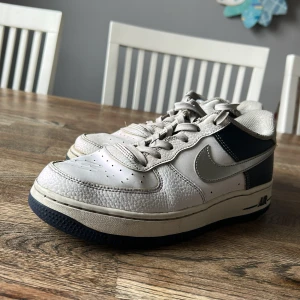 Nike Air Force 1 Low i vitt och mörkblått - Säljer ett par klassiska Nike Air Force 1 Low med vit ovandel i skinn och mörkblå detaljer. Skorna har snörning, rund tå och platt sula. Perfekta för dig som gillar tidlös streetstil. 