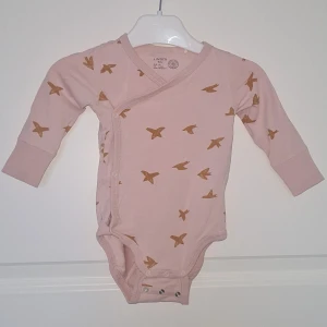 Ljusrosa omlottbody med fågelmönster från Lindex - Supersöt ljusrosa omlottbody från Lindex med extra långa ärmar och dubbel knäppning för extra långbarhet. Bodyn har ett gulligt mönster med små senapsgula fåglar över hela plagget. Perfekt för de minsta!