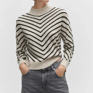 Randig stickad tröja med hög krage - Säljer en beige stickad tröja med svarta ränder i V-mönster och hög krage. Tröjan har långa ärmar och ribbade muddar vid ärmslut och nederkant. Perfekt för dig som gillar en stilren och mysig look.