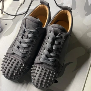 Gråa  Louboutin med spikar  - Säljer ett par gråa sneakers från Christian Louboutin med coola nitar på tån och klassisk röd sula. Skorna har snörning och är tillverkade i skinn med en lyxig känsla. Perfekta för dig som vill sticka ut med din stil. Storlek 38