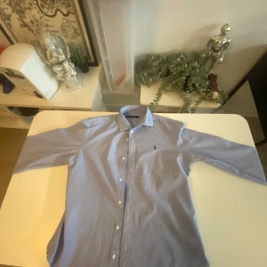 Blå skjorta från Ralph Lauren - Klassisk ljusblå skjorta från Ralph Lauren med broderad logga på bröstet. Skjortan har lång ärm, knappar framtill och en tidlös design som passar till många tillfällen. Perfekt för dig som gillar stilrena plagg med premiumkänsla.